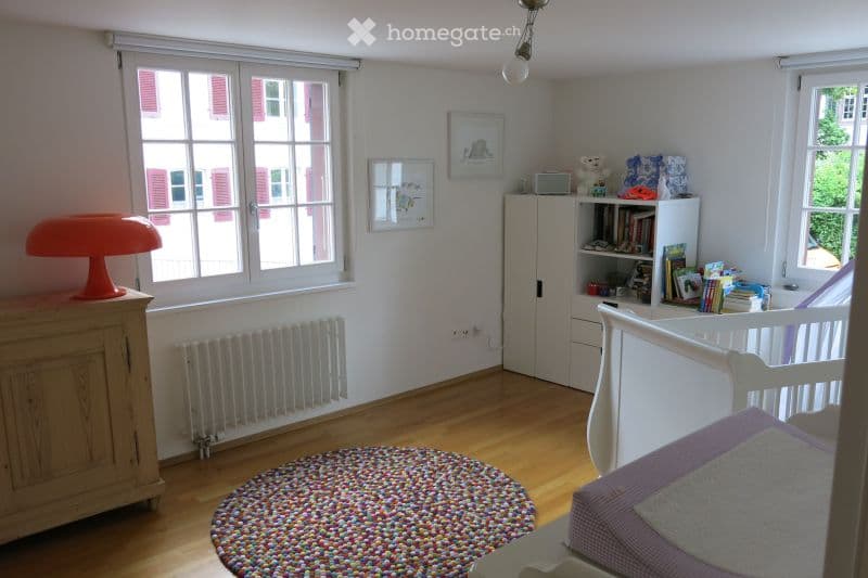 Bright, child-friendly bedroom with wooden flooring, large windows, a white bed with purple bedding, a colorful round rug, a white cabinet, a bookshelf filled with books and toys, and an orange lamp on a wooden dresser.