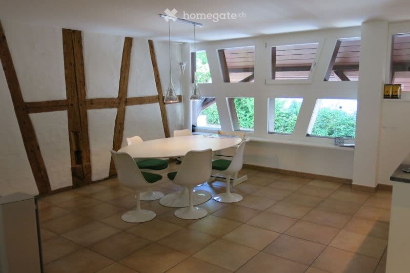 Modern dining room with a white round table, six white chairs, pendant lights, and large windows with wooden shutters.