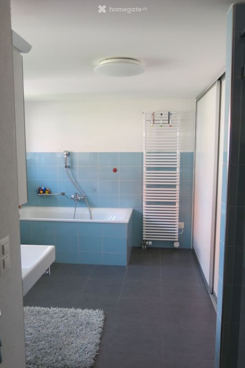 Modern bathroom with blue tiles, a bathtub, a showerhead, a white radiator, a small stool, and a grey rug on the floor.