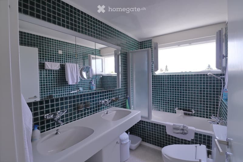Bathroom with green tiled walls, double sink, toilet, bathtub, and large mirror. Towels and toiletries are visible.