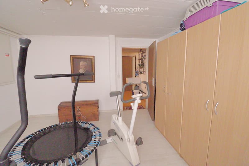 A room with a treadmill, exercise bike, a wooden cabinet, a framed painting, and a small chest. The space is well-lit with white walls and a neutral color palette.