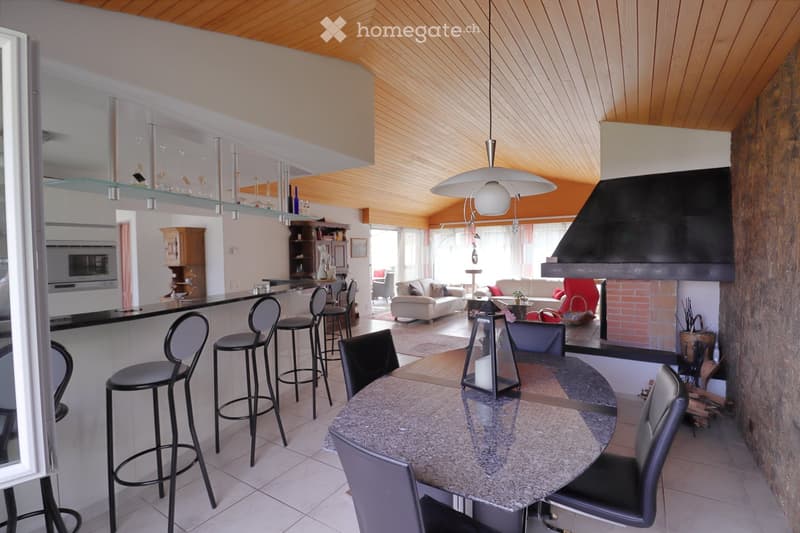Open-plan living and dining area with a modern kitchen, featuring a sofa, dining table with chairs, bar stools, and a fireplace.