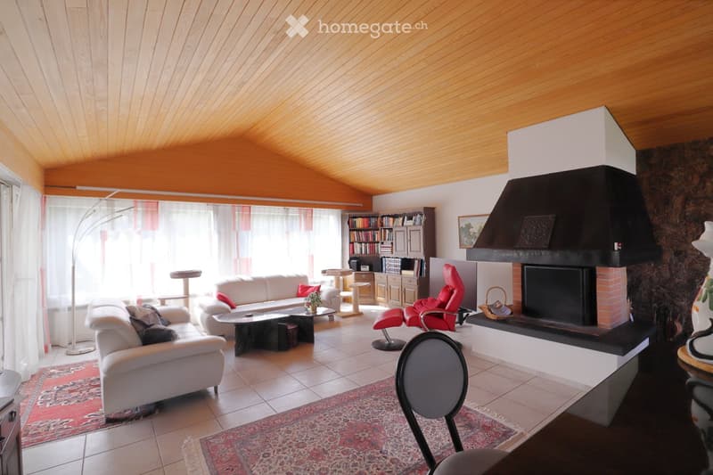Bright living room with wooden ceiling, large windows, white sofa, red chair, coffee table, bookshelf, and a fireplace with a black hood.