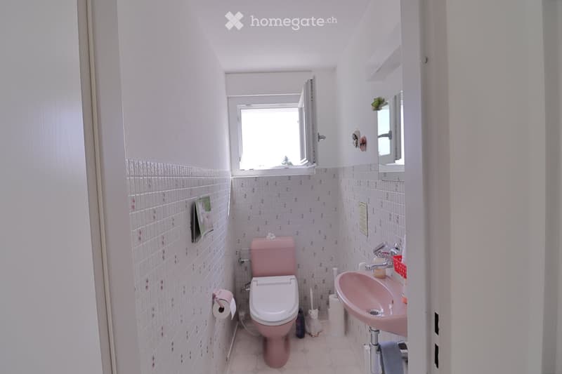 A small bathroom with pink tiles, a pink toilet, a pink sink, a mirror, and a small window. A cat is present near the toilet.