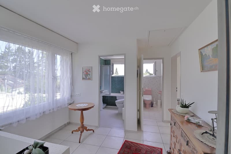 A well-lit hallway with white walls, tiled flooring, and large windows covered with sheer curtains. The hallway features two open doors leading to bathrooms, each with visible fixtures such as sinks, toilets, and bathtubs. There are small wooden tables, p