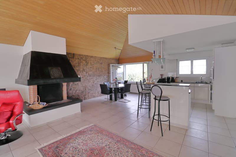 Open-plan living and dining area with a fireplace, red leather sofa, dining table with chairs, bar stools, and a modern kitchen area with a counter and appliances.