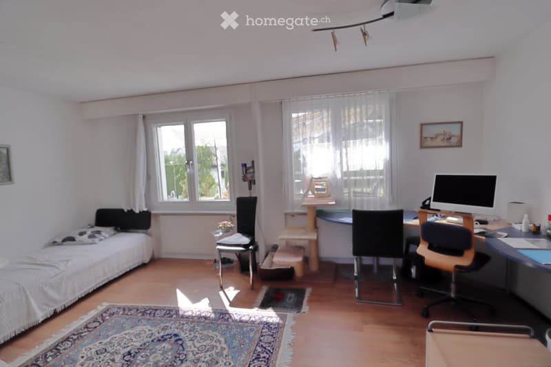 A small, well-lit room with a bed, desk, and chair. The room features wooden flooring, large windows with white curtains, and a colorful rug.
