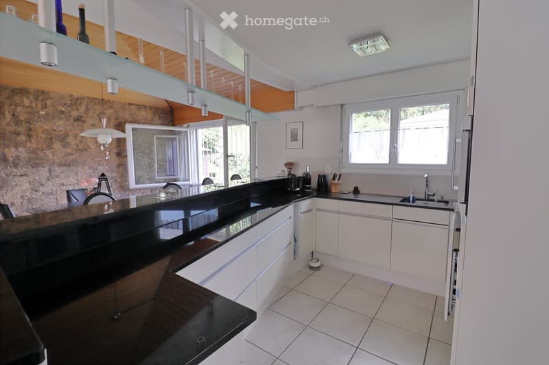 Modern kitchen with white cabinets, black countertops, and a stone accent wall. Features a sink, faucet, coffee maker, and a glass shelf with decorative items.