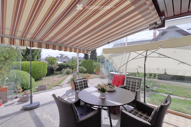 A covered patio with a round table, four wicker chairs, a large umbrella, and a flower pot on the table. The patio has a striped awning and overlooks a garden with plants and trees.