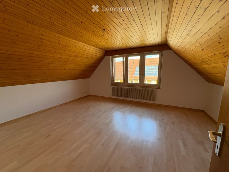 Empty attic room with wooden floors, wooden slanted ceilings, white walls, and a window showing a view of neighboring rooftops.