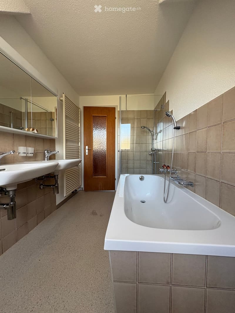 Modern bathroom with a bathtub, two sinks, a large mirror, and a wooden door leading to another room.