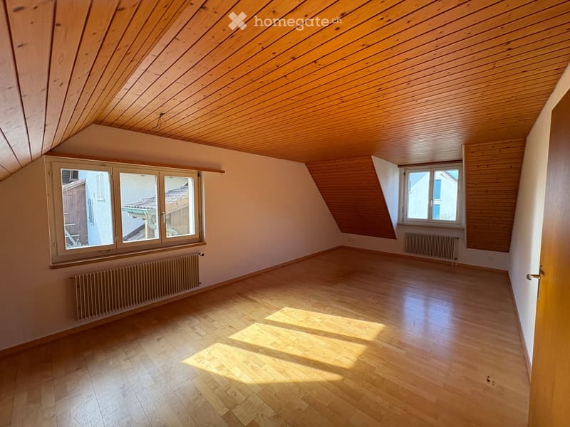 Empty attic room with wooden floors and slanted wooden ceilings, featuring two windows and radiators.