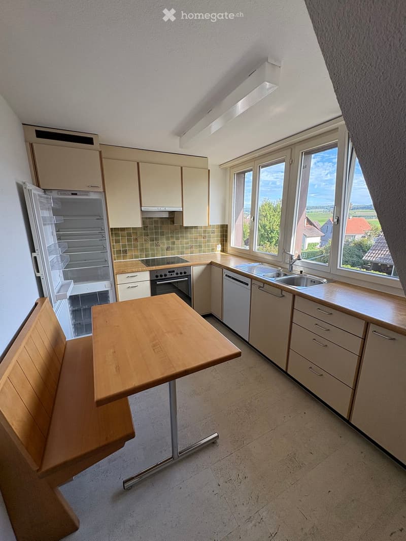 Modern kitchen with light-colored cabinets, stainless steel appliances, and a wooden dining table with bench seating. Large windows offer a view of the outdoors.