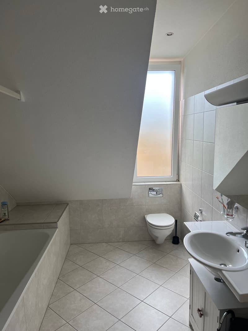 Modern bathroom with a toilet, sink, and bathtub. The room features tiled walls and floor, a window with frosted glass, and a small shelf with a bottle on the left side.