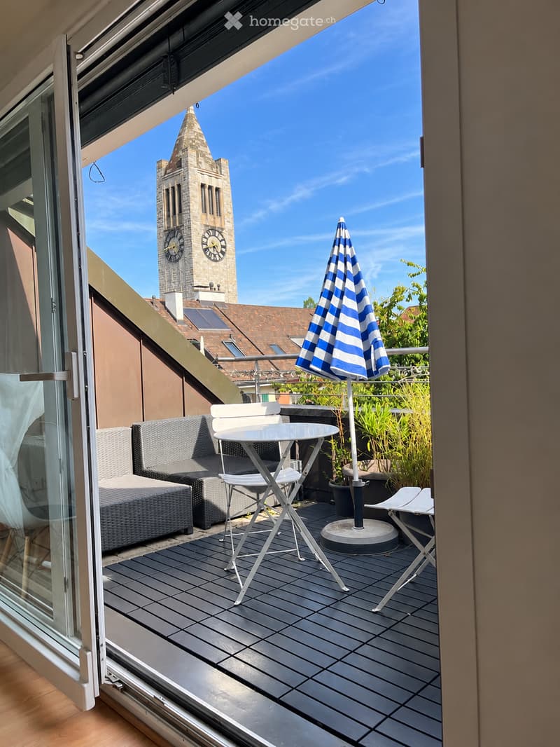 Balcony with wooden decking, grey sofa, white table and chairs, umbrella, and view of a clock tower.