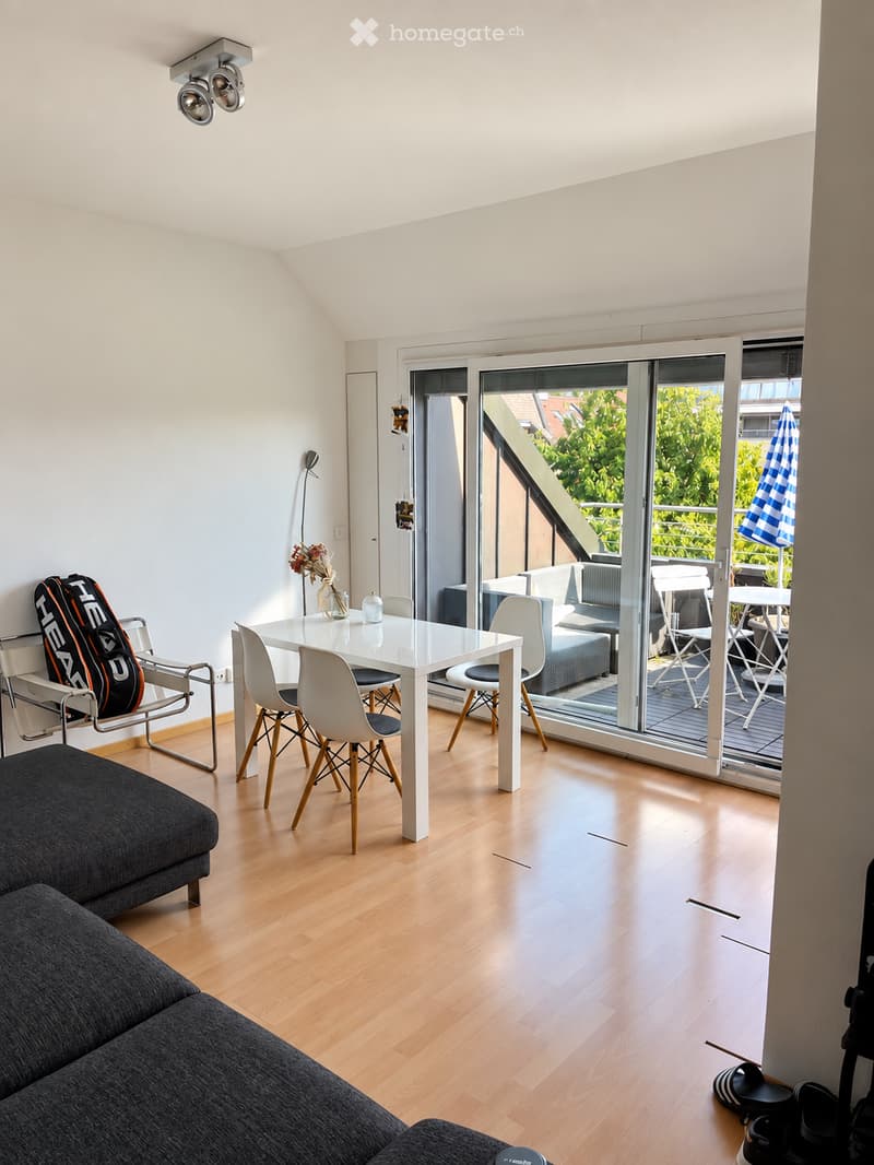 Modern living and dining area with light wooden flooring, white dining table with chairs, grey sofa, and large sliding glass doors leading to a balcony with outdoor furniture.