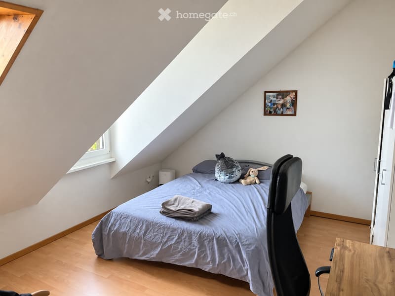 Cozy attic bedroom with slanted ceilings, wooden flooring, a bed with blue bedding, a chair, a small table, and decorative items including a picture frame and toys.