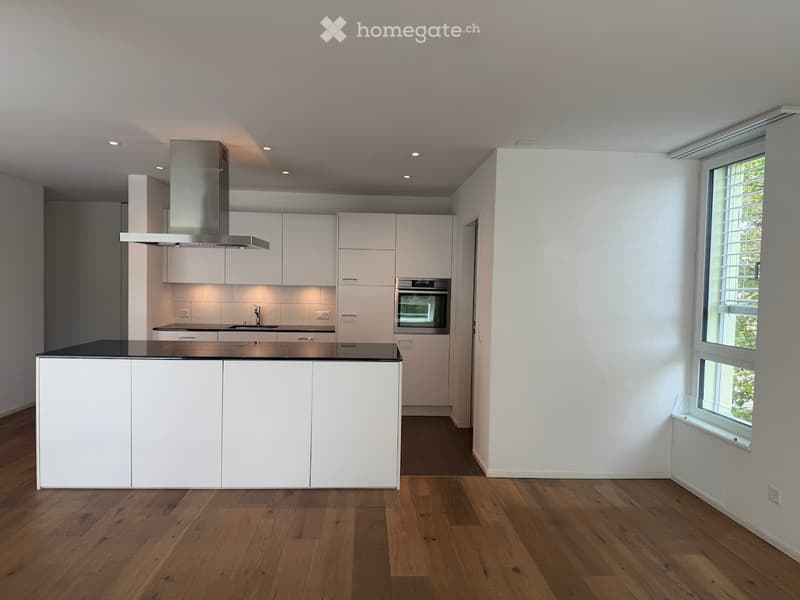 Modern, minimalist kitchen with white cabinets, a central island, and integrated appliances including an oven and range hood. The space features wooden flooring and large windows allowing natural light.