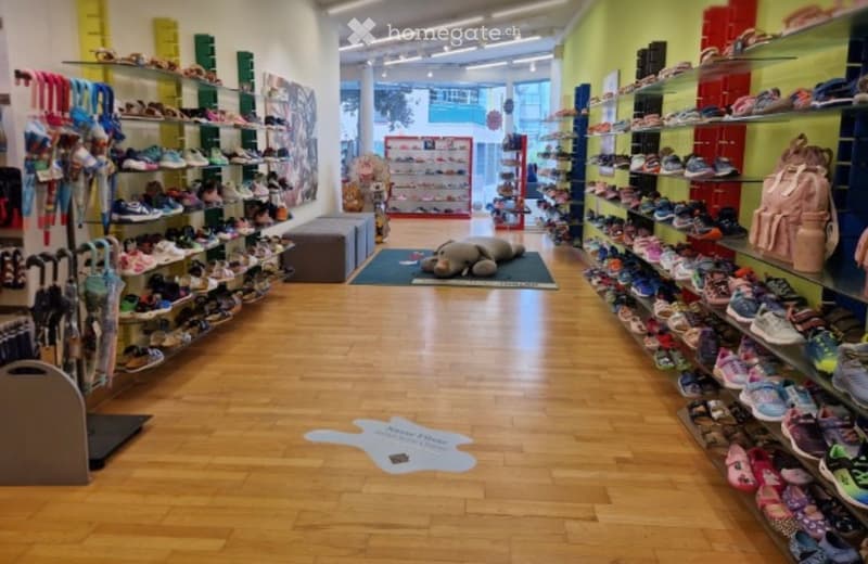 A brightly lit shoe store with wooden flooring, shelves filled with various shoes, a plush toy on the floor, and a backpack on display.