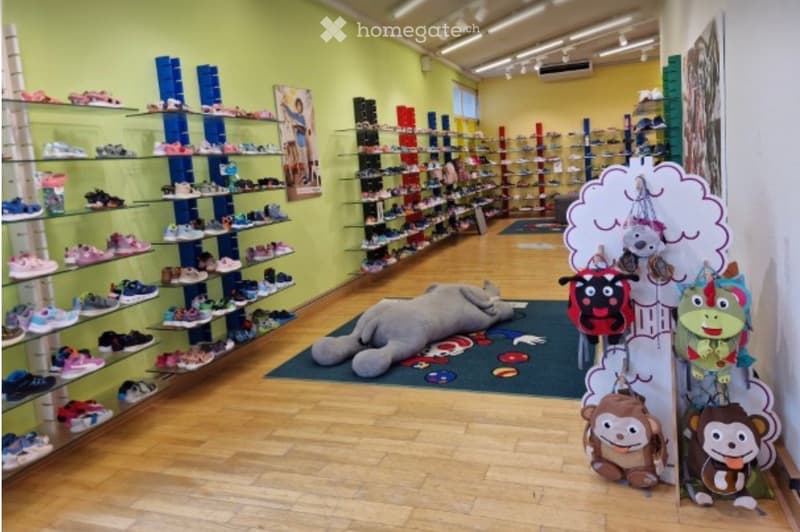 A brightly lit shoe store with multiple shelves displaying various shoes, a large plush toy on the floor, and a colorful display with hanging toys.