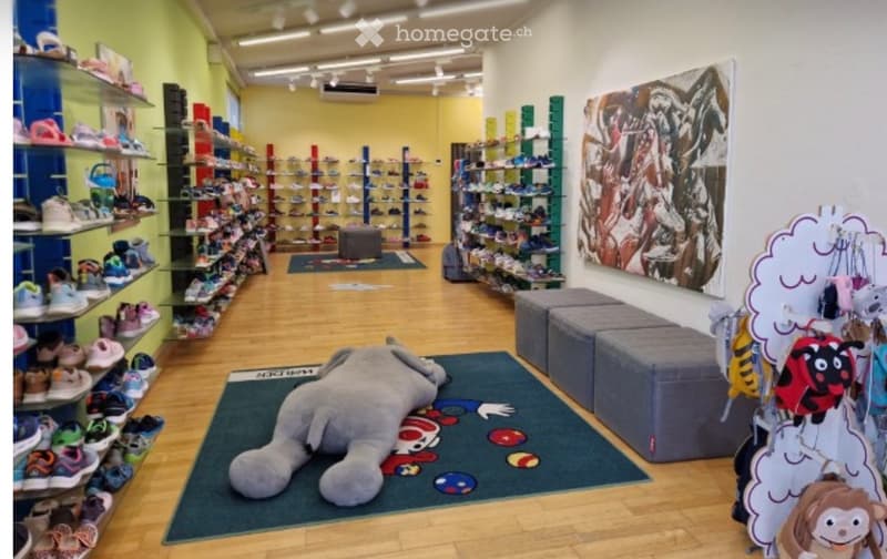 A brightly lit children's shoe store with wooden flooring, shelves filled with shoes, a large plush toy, and colorful decor including a cloud-shaped display and a large picture on the wall.