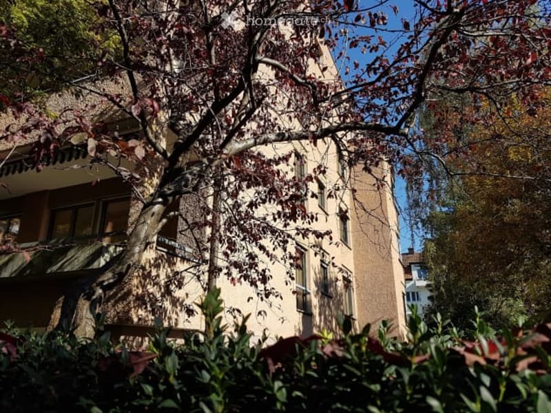 A multi-story residential building with a beige facade, multiple windows, and a tree partially obscuring the view.
