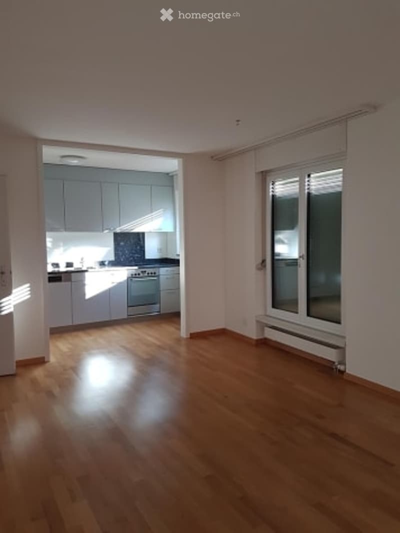 Empty room with wooden flooring, large window with blinds, and an adjacent kitchen area with light-colored cabinets and stainless steel appliances.