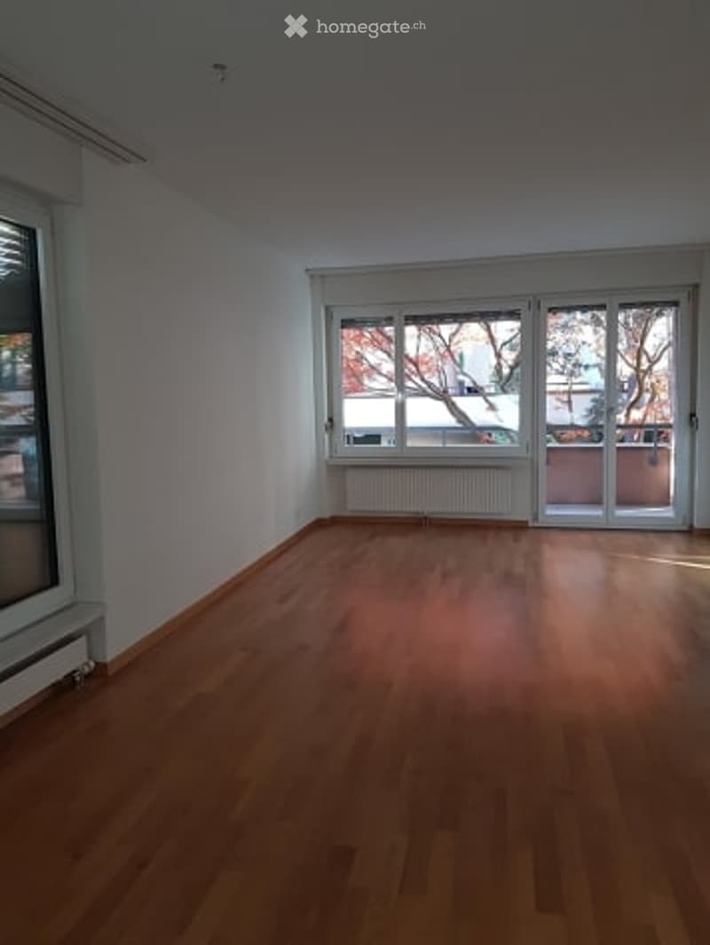 Empty room with wooden flooring, white walls, large windows, and sliding glass doors leading to a balcony.