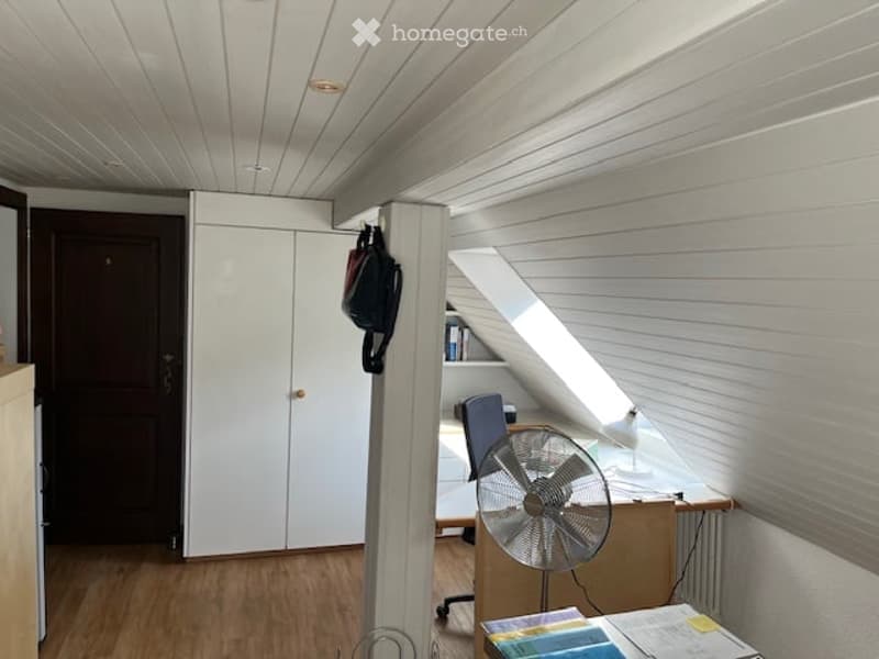 Bright room with wooden flooring, white walls, and a slanted ceiling. Features a desk with a chair, a fan, a cabinet, and a backpack hanging on the wall.