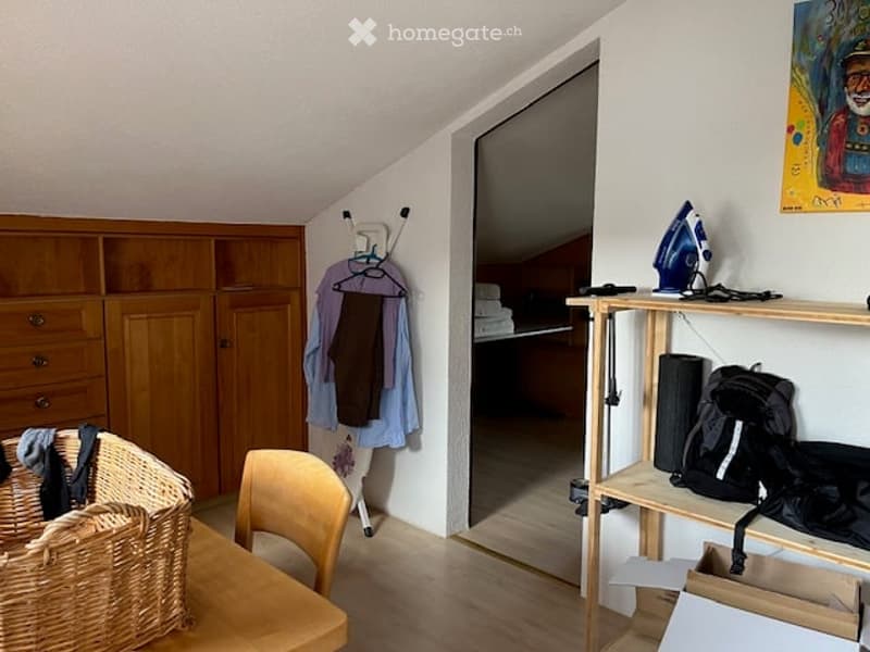 A cozy, slanted-ceiling room with a wooden table, chair, cabinet, shelf, and various personal items including a backpack and an iron.