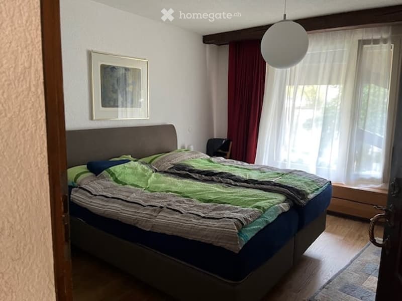 Cozy bedroom with a double bed, green and grey bedding, a nightstand, a chair, and a picture frame on the wall.