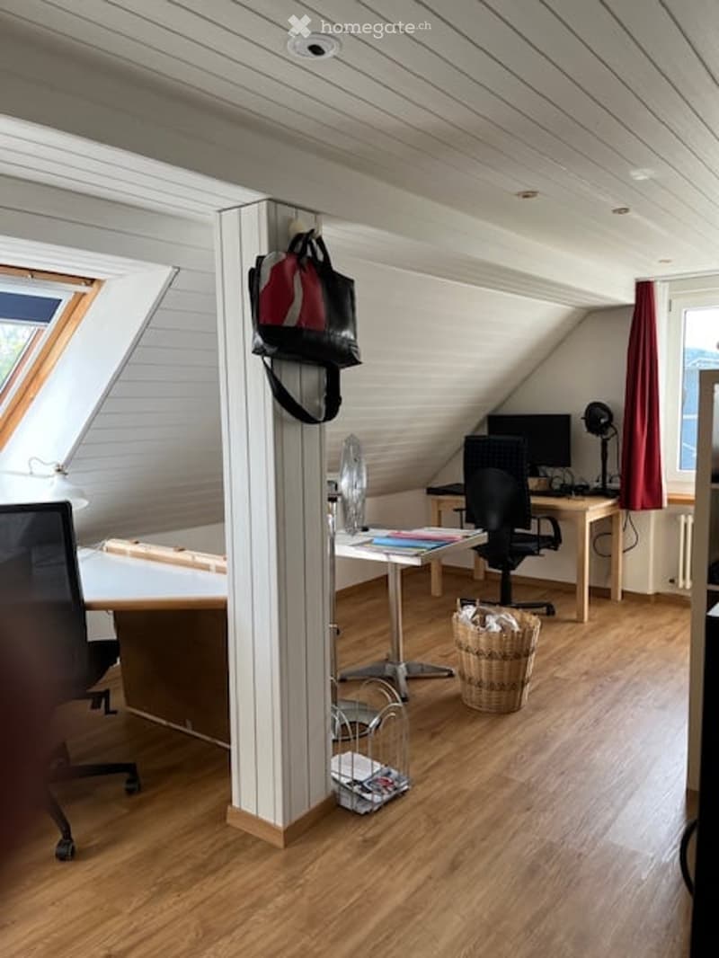 Attic room with wooden flooring, slanted white ceiling, large window, desk with chair, computer monitor, lamp, curtain, wicker basket, and handbag hanging on a stand.