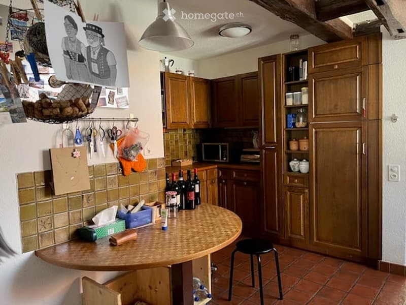 A cozy kitchen with wooden cabinets, a round wooden table, a black stool, and various kitchen items including wine bottles and tools on the wall.