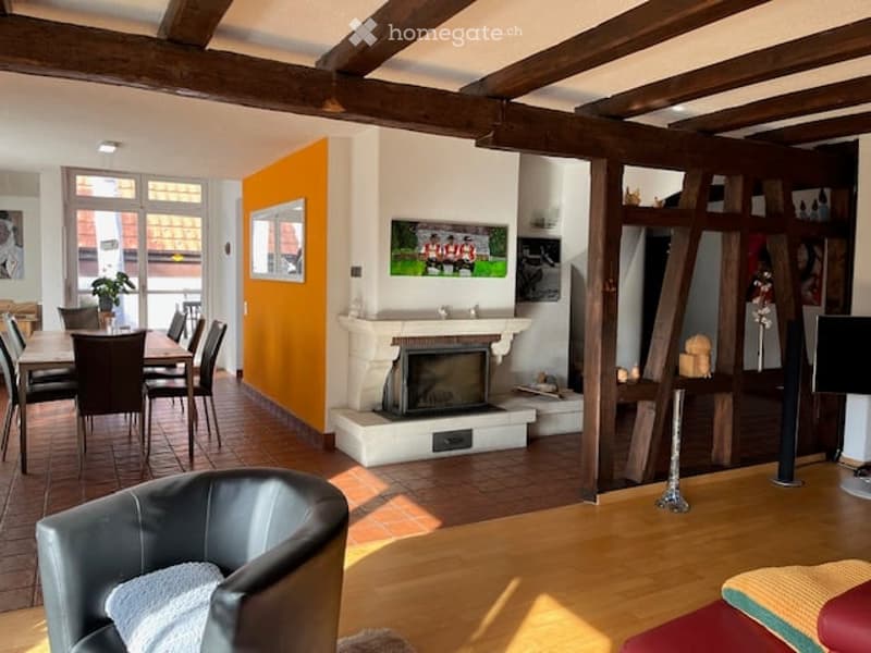 Living room with exposed wooden beams, fireplace, large windows, dining area, leather armchair, and TV.