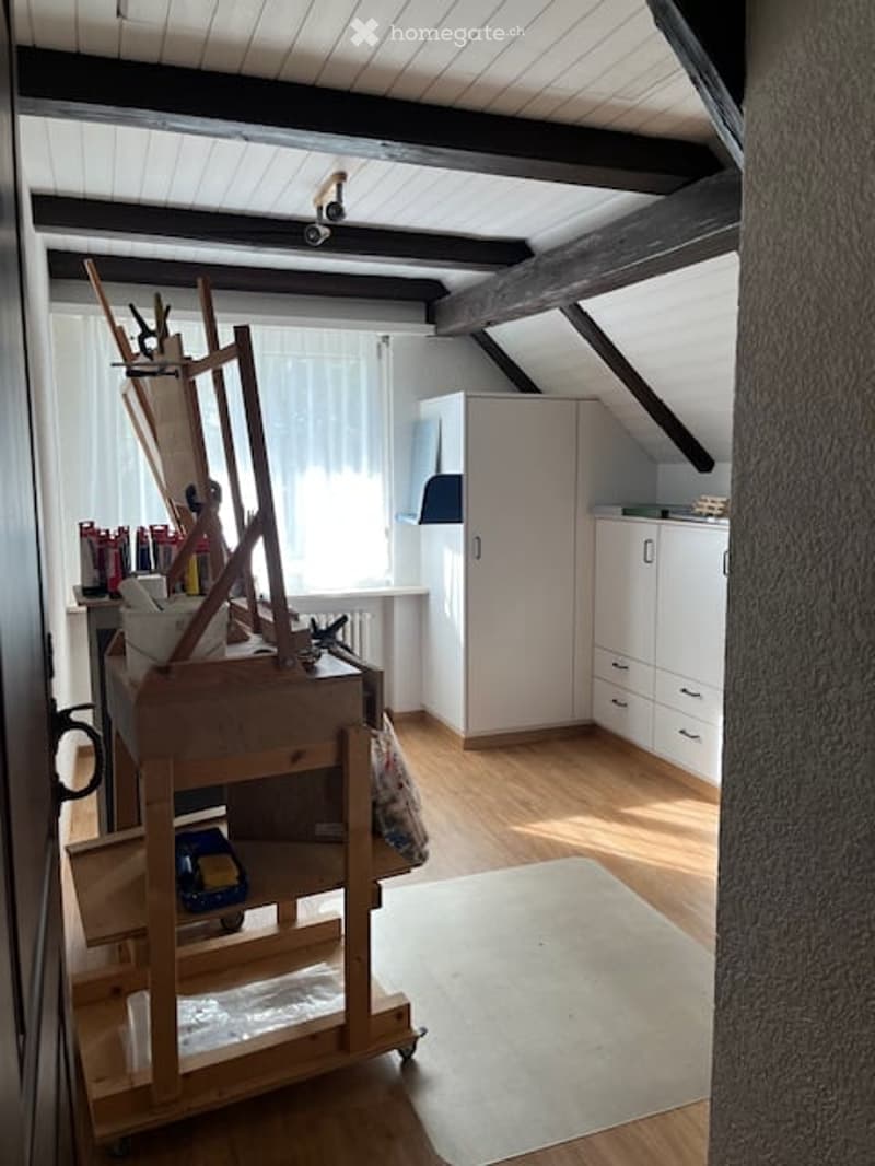 A well-lit room with wooden floors and white walls, featuring a large easel, a cabinet, a small table with art supplies, and a toy car on a shelf.