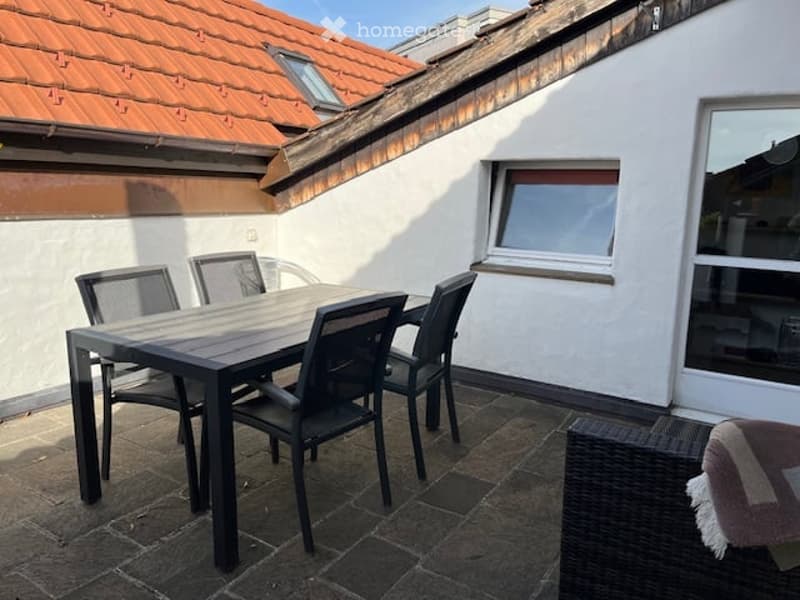 Balcony with a wooden dining table and four black chairs, a wicker chair with a cushion, and a window with blinds.