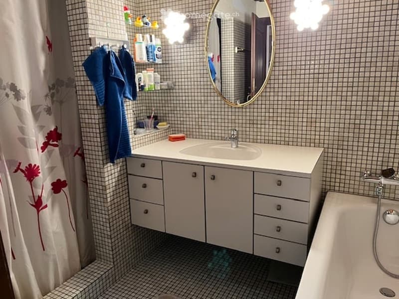Modern bathroom with white cabinetry, a sink, a mirror, a bathtub, and a showerhead. The walls are tiled with small square tiles, and there is a towel hanging on a rack.