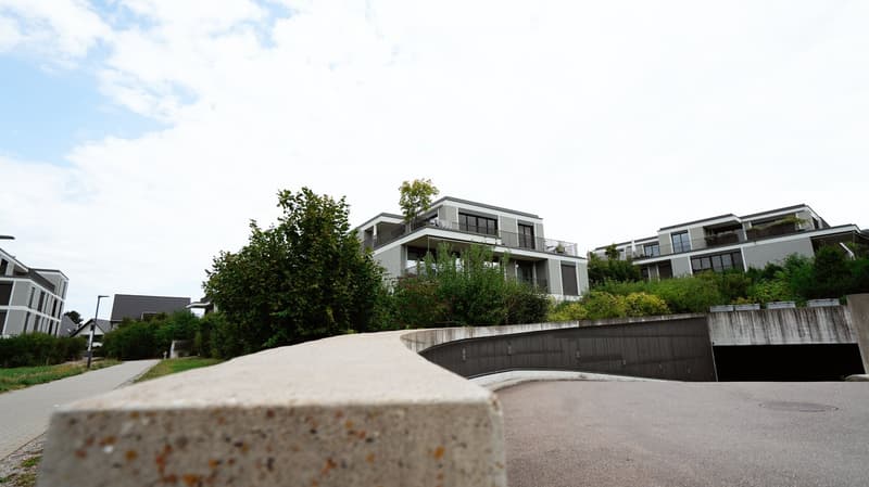 Modern multi-story residential building with balconies, surrounded by greenery and a paved driveway leading to a garage.