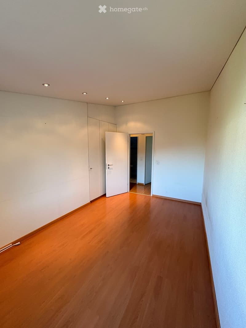 Empty room with wooden flooring, white walls, and recessed ceiling lights. Features an open closet and a doorway leading to another room.