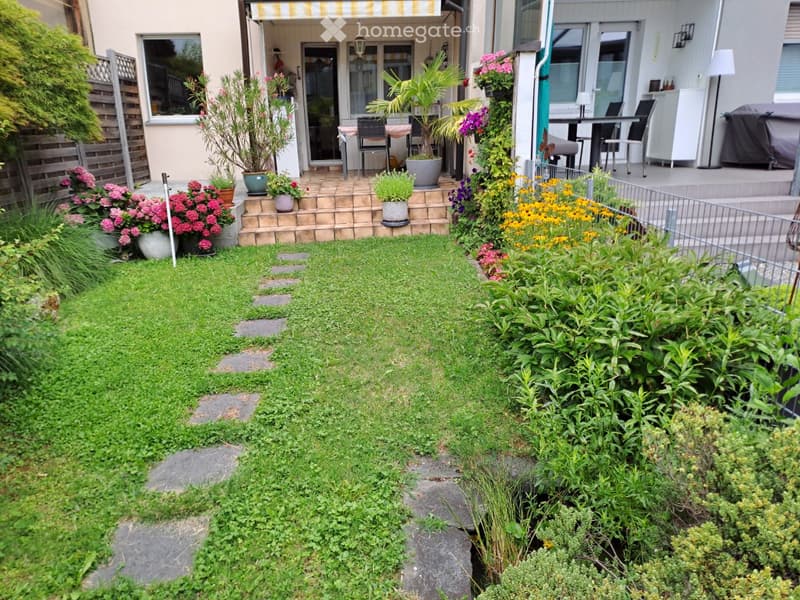 Small garden with a stone pathway, green grass, potted plants, and a patio area with outdoor furniture.