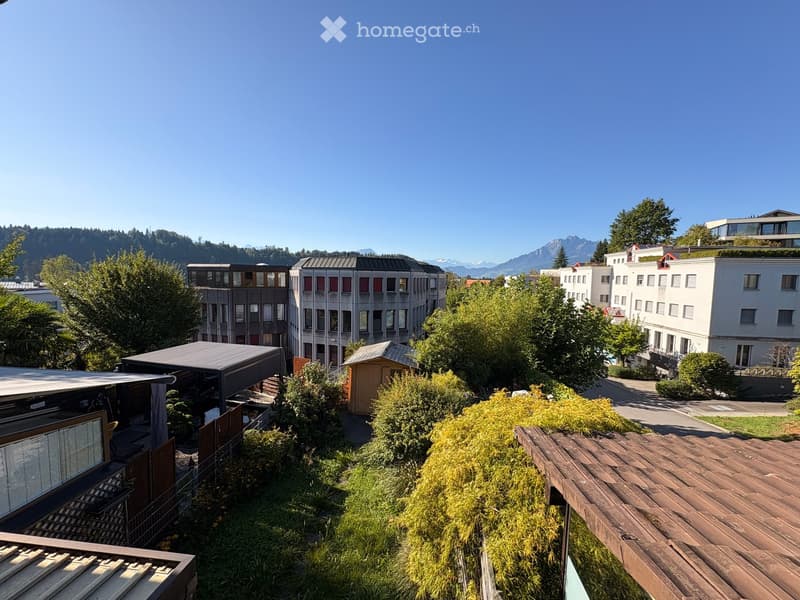 A panoramic view of a modern residential area with multiple buildings, greenery, and a clear blue sky. The scene includes a mix of contemporary architecture, trees, and a distant mountain range.
