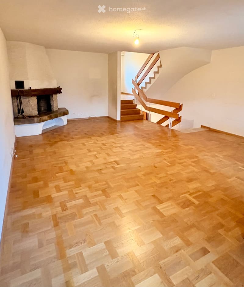 Empty room with wooden flooring, white walls, and a staircase leading upwards. A fireplace is visible on the left side.