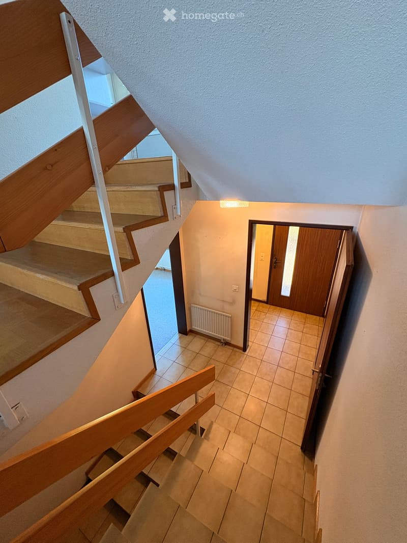 Wooden staircase with white railings leads to a hallway with tiled flooring and a wooden door at the end.
