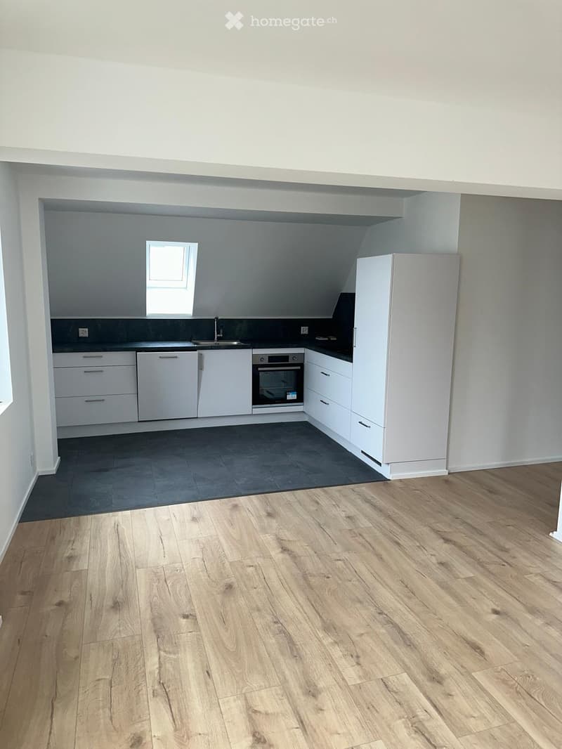 Modern kitchen with white cabinets, stainless steel appliances, and a dark countertop. The floor is light wood, and there is a small window above the sink.