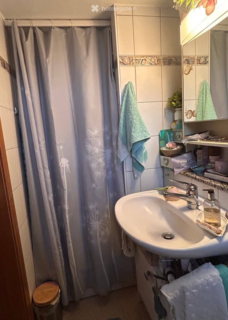 Bathroom with a sink, mirror, towel, shower curtain, and a small stool. The sink has a modern faucet and a soap dispenser. A towel hangs on the wall next to the shower area.