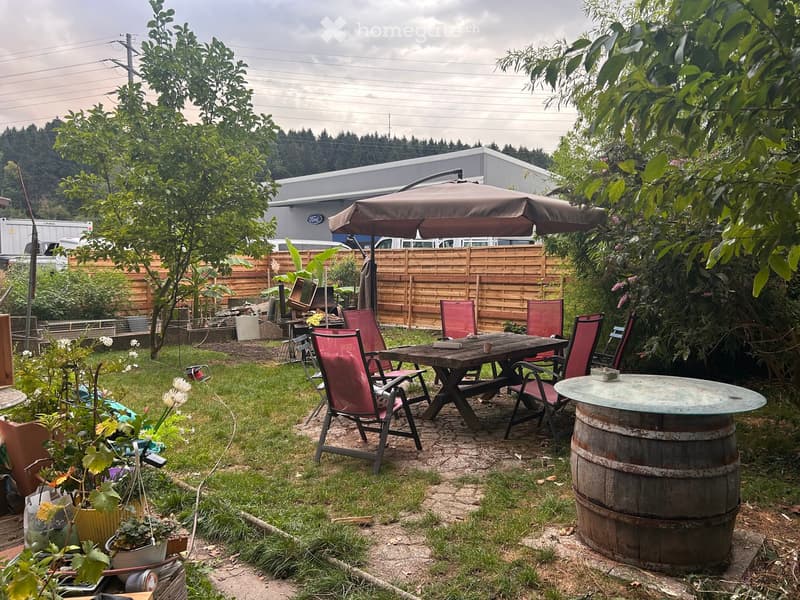 Outdoor patio area with a wooden picnic table, red chairs, umbrella, and surrounding greenery.