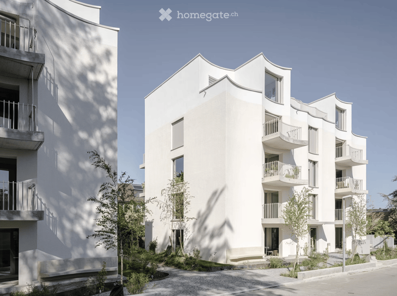 Modern white apartment building with multiple balconies and contemporary design, surrounded by small trees and greenery.