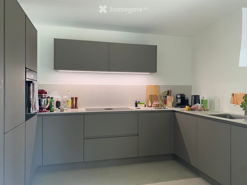 Modern kitchen with grey cabinets, stainless steel appliances, and a white countertop. Visible items include a mixer, coffee maker, cutting board, and various kitchen utensils.