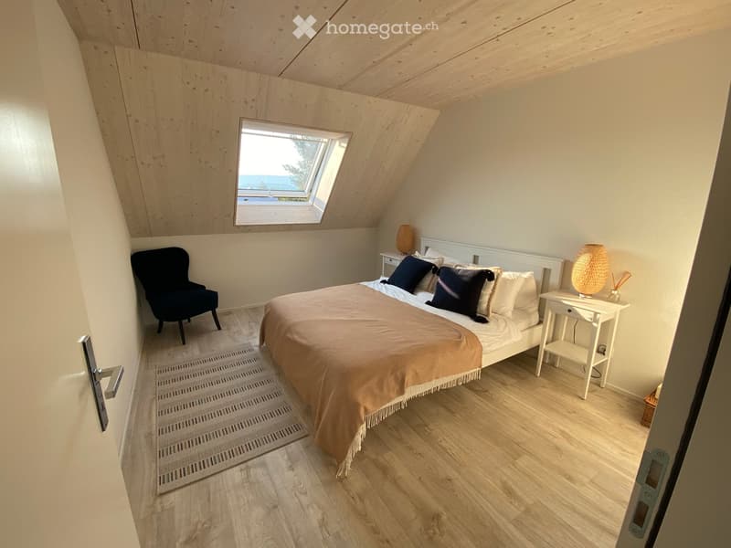 Cozy attic bedroom with wooden walls and ceiling, skylight window, bed with pillows and blanket, nightstand with lamp, and a small black armchair.