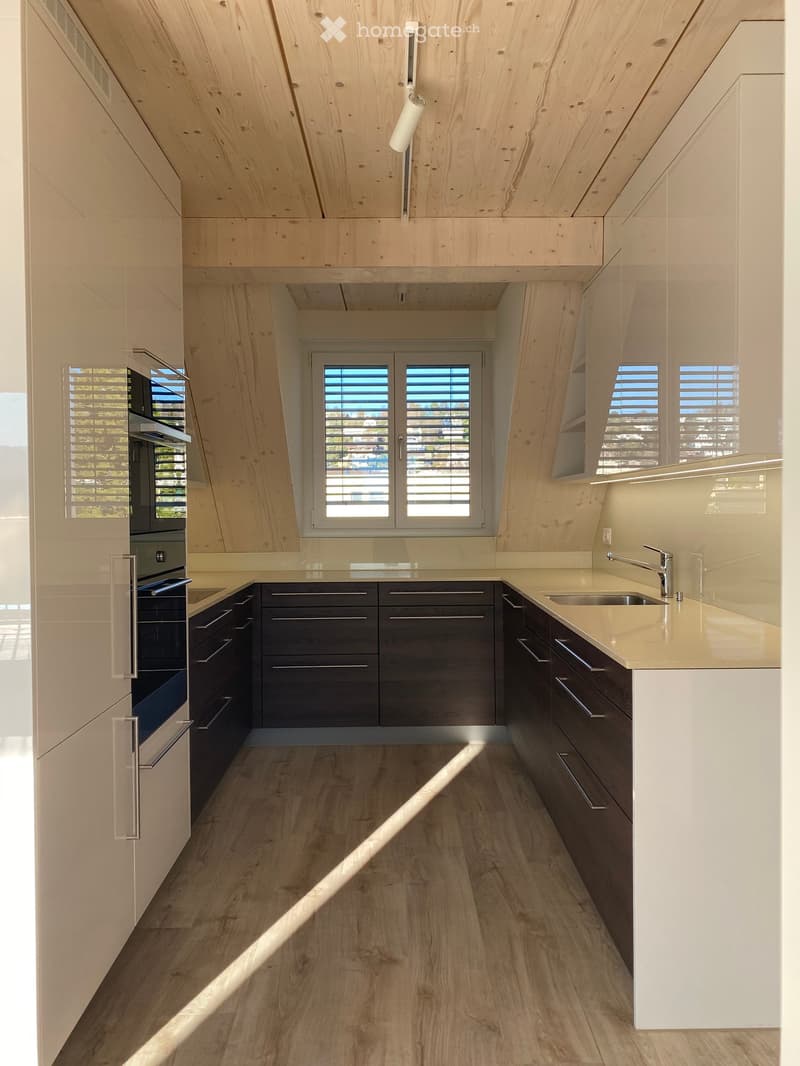 Modern kitchen with light wood cabinets, dark lower cabinets, a central island, and a sleek stainless steel oven. Natural light streams in from large windows with blinds.